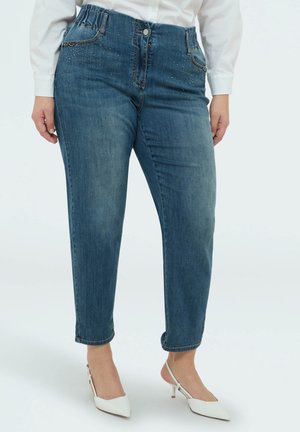 Pantalones vaqueros de mezclilla azul para mujer con cintura elástica y pequeños detalles de pedrería, combinados con tacones blancos tipo slingback.