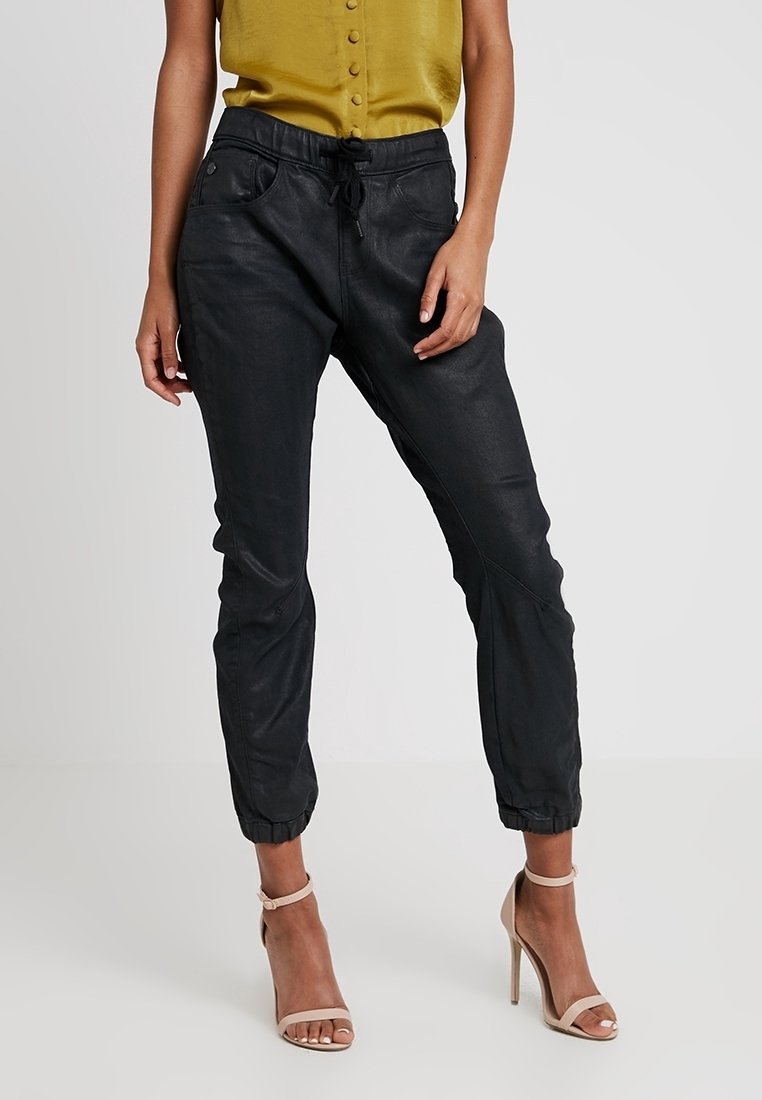 Svarta joggerbyxor med dragsko i midjan, smalnade ben och svag glans; kombinerat med en senapsgul blus och hudfärgade högklackade sandaler.