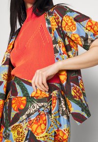 Haut rouge à côtes associé à un kimono à motifs floraux colorés. Les couleurs vives incluent l'orange, le bleu et le vert. Le tissu semble léger et texturé.