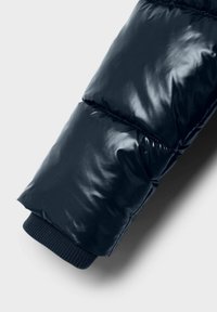 Manche d'une veste matelassée noire brillante avec des coutures visibles et un poignet élastique à côtes sur fond blanc.