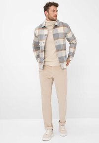 Uomo che indossa un dolcevita beige, pantaloni in corduroy beige, sneakers bianche e una giacca a quadri grigia, bianca e beige, mentre sta in piedi con le mani in tasca.