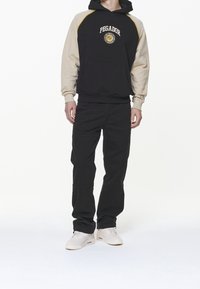 Felpa con cappuccio nero e beige con logo, tasca a marsupio, polsini a costine; abbinata a pantaloni neri e sneakers bianche per un look casual.