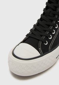 Baskets en toile noires avec embout et semelle en caoutchouc blanc, présentant un profil de semelle texturé et des lacets noirs, rehaussés de détails de couture blancs.