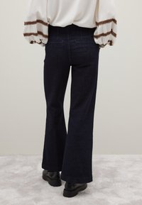 Dunkle Denim-Flared-Hosen mit hoher Taille und zwei Gesäßtaschen, kombiniert mit einem weißen Oberteil mit brauner Einfassung an den Ärmeln.