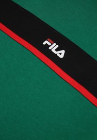 Tessuto verde con una fascia a strisce nera e rossa che presenta il logo "FILA" in bianco. Il materiale è morbido e leggermente strutturato.