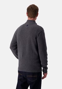 Maglione grigio lavorato a maglia con colletto alto, polsini e orlo a coste, con una piccola etichetta logo sul retro. Indossato con jeans scuri.