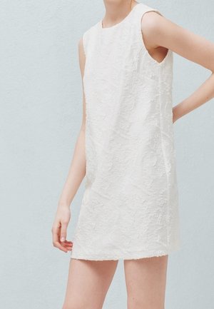 Mini-robe blanche texturée sans manches portée par une personne debout contre un mur bleu clair, une main détendue le long du corps et l'autre sur la hanche.