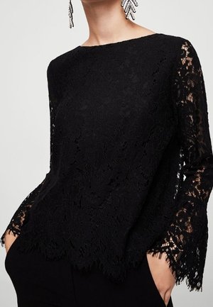 Blouse noire à manches longues en dentelle avec motifs floraux et bords festonnés, portée avec un pantalon noir, mains dans les poches, et boucles d'oreilles remarquables visibles.