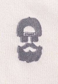 Beige Stoff mit einem dunkelgrauen gestickten Design, das ein stilisiertes Totenkopf-Motiv mit Schnurrbart zeigt. Das Motiv ist zentriert, hat einen abgerundeten oberen Teil und detaillierte Gesichtszüge.