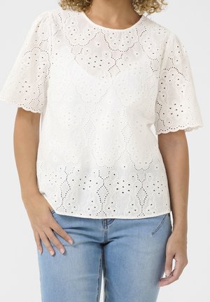 Hvid bluse med korte ærmer med hulbroderi og bølgede kanter, båret med lyseblå jeans, hånd hvilende på hoften.