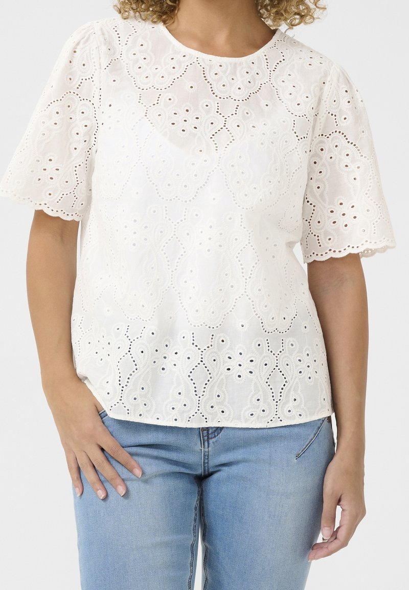 Blouse blanche à manches courtes avec broderie anglaise et bords festonnés portée avec un jean bleu clair, main reposant sur la hanche.