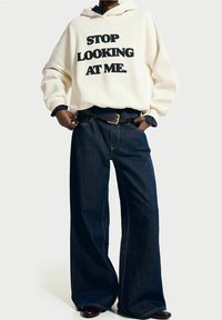 Kávově krémová mikina s kapucí a výrazným černým textem "PŘESTAŇTE SE NA MĚ DÍVAT", doplněná tmavě modrými širokými džíny. Hnědý opasek a boty.