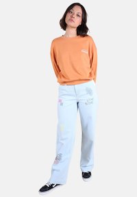 Chemise orange à manches longues avec logo ; jean large bleu clair avec motifs brodés colorés ; baskets noires avec semelles blanches.