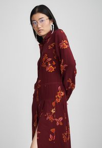 Βουργουνδί φόρεμα με κεντήματα λουλουδιών σε πορτοκαλί και κίτρινο. Διαθέτει μακριά μανίκια, κλείσιμο με κουμπιά, πλευρική σχισμή και χαλαρή εφαρμογή.