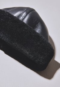 Juoda odinė žieminė kepurė su dirbtinio kailio kailiuku ir storu puriu juostos apvadu, atvaizduota iš arti ant šviesaus paviršiaus.