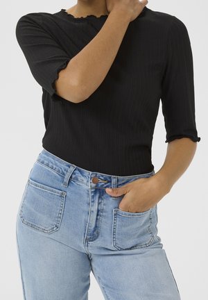 Osoba ubrana w czarną prążkowaną bluzkę z falbankami na krawędziach i jasnoniebieskie, wysokowciskowe dżinsy, z jedną ręką w kieszeni, a drugą dotykającą szyi.