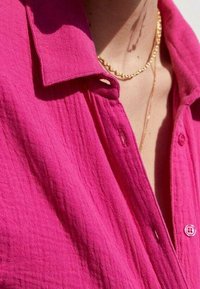 Chemise fuchsia à boutons avec un tissu texturé et froissé, dotée d'un col. Un collier en fine chaîne d'or est visible autour du col.