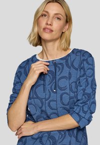 Blauer strukturierter Pullover mit einem wellenförmigen Muster, Rundhalsausschnitt mit weißem Besatz und verstellbarem Kordelzug. Dreiviertelärmel und lockere Passform.