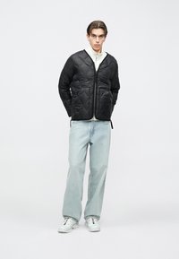 Černá prošívaná bunda se zipem vpředu, nošená přes bílé tričko, v kombinaci s světlými modrými džíny a bílými teniskami. Jednoduchý a čistý design.