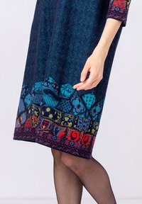 Haljina do koljena u tamnoplavoj boji s teksturiranim materijalom, koja sadrži šareni pojas s geometrijskim oblicima i cvjetnim uzorcima. Dugi rukavi.