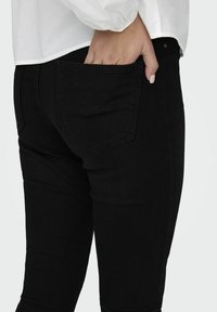 Jeans de mezclilla negra con un diseño ajustado. Detalle notable en el bolsillo trasero y textura suave. Llevados con una camisa blanca de manga larga.