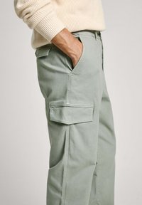 Svetlozeleni cargo hlače z bočnimi žepi in žepom za nalepke, izdelane iz mehke in zračne tkanine, s sproščenim krojem in šitimi detajli.