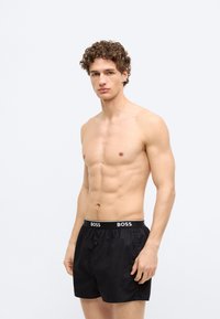 Čierne boxerky s hladkou textúrou a čiernym pásom s logom "BOSS". Navrhnuté s gombíkovým zapínaním vpredu.