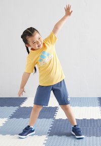 Keltainen puuvillapaita, jossa on sininen Adidas-logo; tummansiniset shortsit; tummansiniset kengät vaaleansinisillä yksityiskohdilla; lapsi venyttelee teksturoiduilla laattalattioilla.