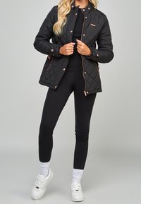Chaqueta negra acolchada con detalles en oro rosa, botones a presión y bolsillos laterales, combinada con leggings negros y zapatillas blancas.