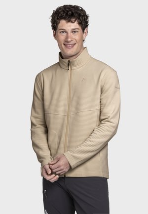 Junger Mann mit lockigem Haar, der lächelt, trägt eine beige Reißverschlussjacke und schwarze Hose, steht vor einem einfachen hellen Hintergrund.