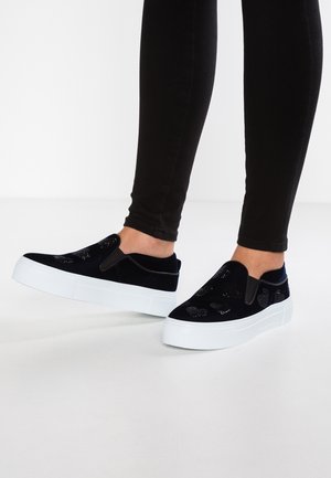 Fødder iført sorte slip-on sneakers med hvide platformsåler og hjerteformede udskæringsdesigns, kombineret med sorte tætsiddende bukser.