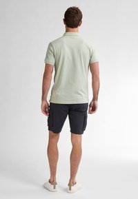 Polo verde chiaro con un motivo testurizzato, abbinato a pantaloni cargo scuri e scarpe bianche casual. Vista da dietro.