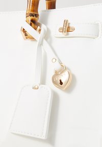 Witte handtas met een bamboehandle, gouden hardware, een hartvormig slot en een bijpassend label met stiksels.