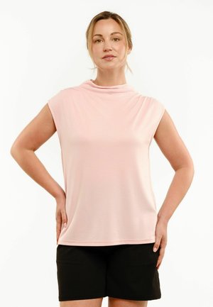 Kobieta z jasnobrązowymi włosami związanymi do tyłu, ubrana w bladoróżowy bezrękawnik i czarne szorty, stoi z rękami na biodrach.