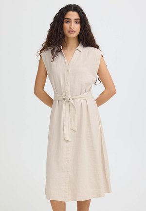 Donna con lunghi capelli ricci che indossa un vestito midi beige senza maniche con bottoni e cintura annodata in vita, in piedi con le mani dietro la schiena.