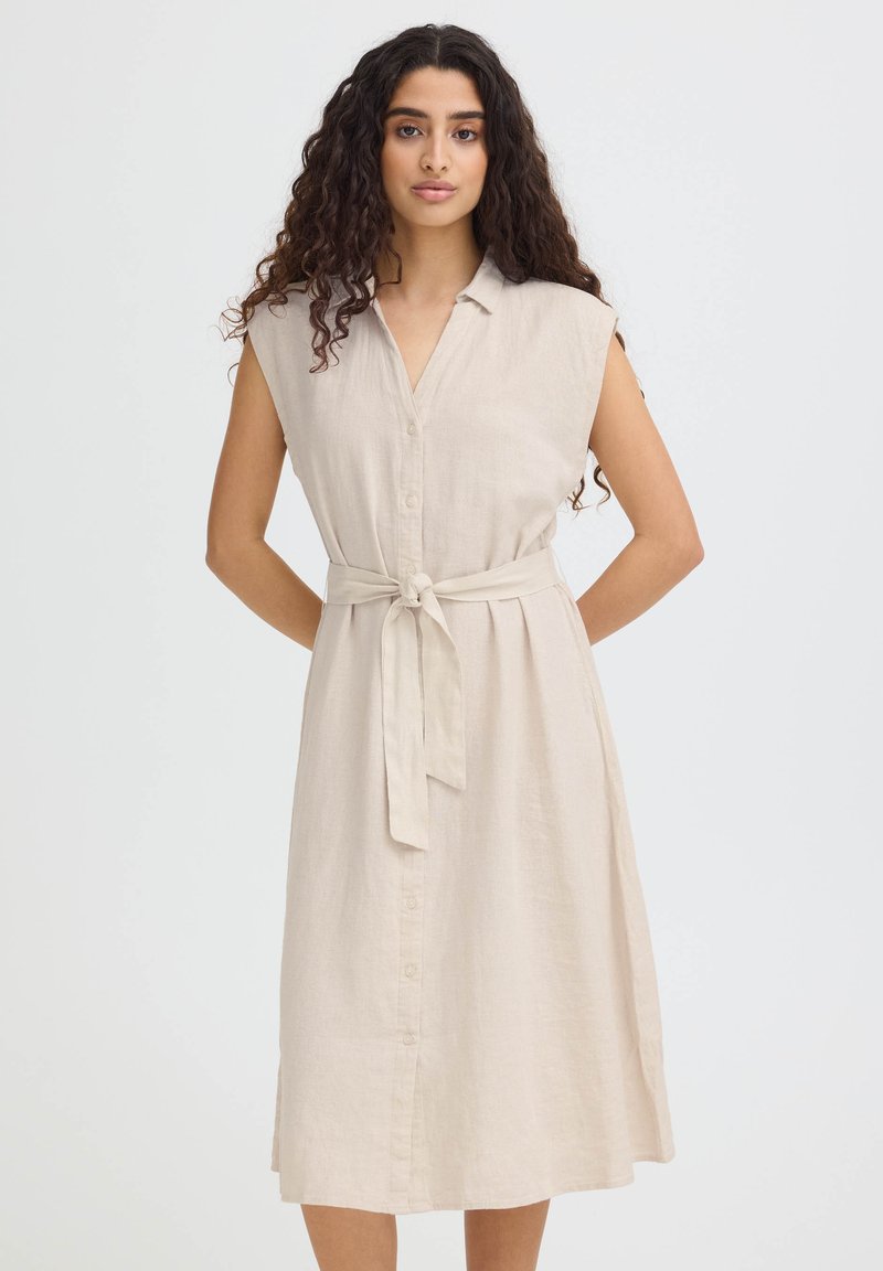 Femme aux longs cheveux bouclés portant une robe midi beige sans manches à boutons avec une ceinture nouée à la taille, debout les mains dans le dos.