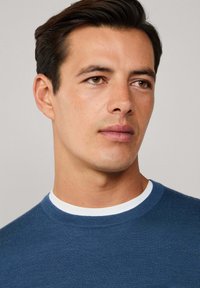 Blauer Strickpullover mit rundem Halsausschnitt und weißem Innenkragen. Glatte Textur, enganliegendes Design und minimalistische Details.