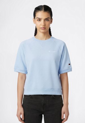 Frau mit geflochtenen Haaren, die ein hellblaues Sweatshirt mit kurzen Ärmeln und Champion-Logos trägt, kombiniert mit schwarzen Jeans vor weißem Hintergrund.