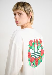 Kremasti oversized dukserica s vibrantnim cvjetnim grafikonom u središtu koji okružuje ikonski Adidas logo, sa crvenim ružama i zelenim lišćem.
