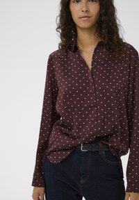 Bluză burgundă cu buline roz deschis, cu nasturi și guler, realizată dintr-un material neted, asortată cu blugi închiși la culoare și o curea neagră.