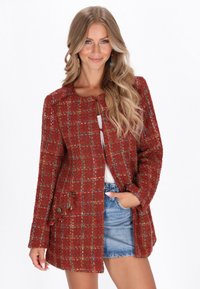 Giacca in tweed rossa con motivo a quadri, scollatura tonda, due tasche frontali e bottoni metallici, abbinata a una maglietta bianca e pantaloni di jeans corti.