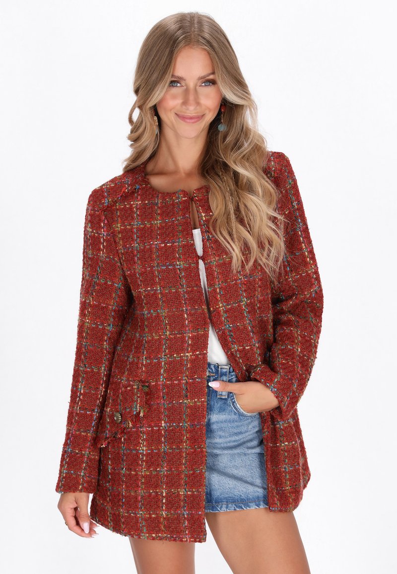 Giacca in tweed rossa con motivo a quadri, scollatura tonda, due tasche frontali e bottoni metallici, abbinata a una maglietta bianca e pantaloni di jeans corti.