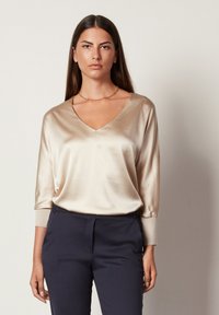 Beige satin topp med V-ringning, långa ärmar och ribbade manschetter, i kombination med mörka skräddarsydda byxor. Slät textur och draperad passform.