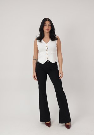 Kvinde stående iført en hvid knapvest, sorte viddebukser og burgunderfarvede spidse sko mod en ensartet lys baggrund.