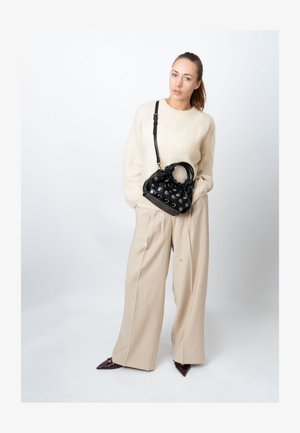 Black leather handbag with metallic studs, rounded handles, and adjustable strap. Model wears a cream sweater and beige wide-leg trousers.