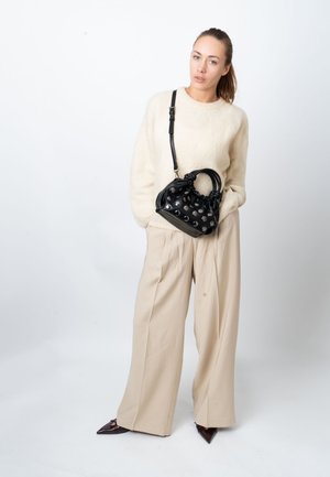 Black leather handbag with metallic studs, rounded handles, and adjustable strap. Model wears a cream sweater and beige wide-leg trousers.