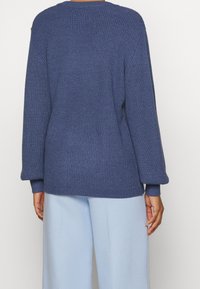 Pull bleu tricoté avec un motif texturé, coupe ample et poignets côtelés. Associé à un pantalon bleu clair, affichant un style décontracté.