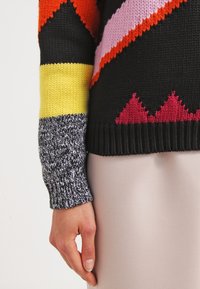 Bunt gestrickter Pullover mit auffälligen geometrischen Mustern in Schwarz, Orange, Pink und Gelb, mit einem strukturierten grauen Ärmel.