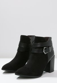 Botas de tobillo de ante negro con punta afilada, que presentan un cierre lateral con cremallera, dos correas de cuero y un detalle de hebilla cuadrada en el tobillo.