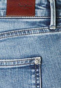 Jeans de mezclilla azul claro con un parche de cuero marrón con "Pepe Jeans" en relieve, un botón metálico y detalles de costura visibles.